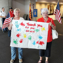 Shelia Parks (left) and Karen Woodrow hold up a banner thanking World War II veterans for their service. The banner was made by Sheila and Drew Parks’ four year old granddaughter’s Montessori preschool class in Lake Forest, California; photo by John McGee.